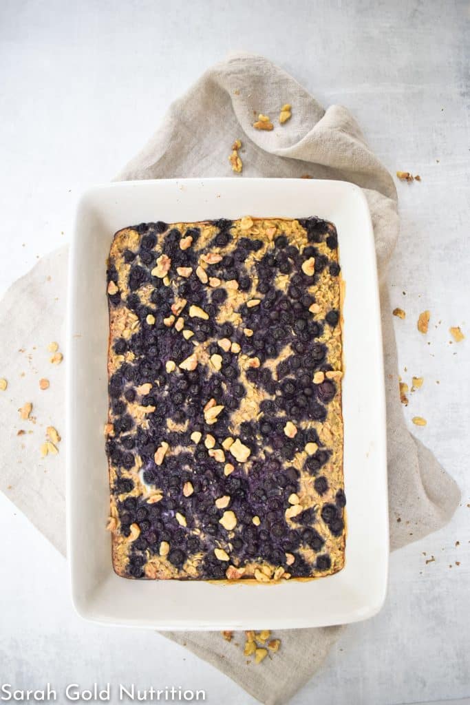 blueberry and walnut high protein baked oatmeal in white dish on top of tan cloth napkin on grey background with walnuts scattered around the dish. 