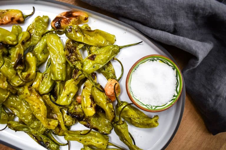 grilled shishito peppers on white serving tray with bowl of sea salt and grey napkin