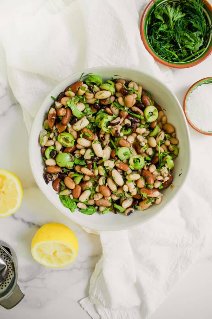 overhead shot of easy bean salad with olives and herbs in large white bowl with lemons and bowl of herbs and salt on marble background 